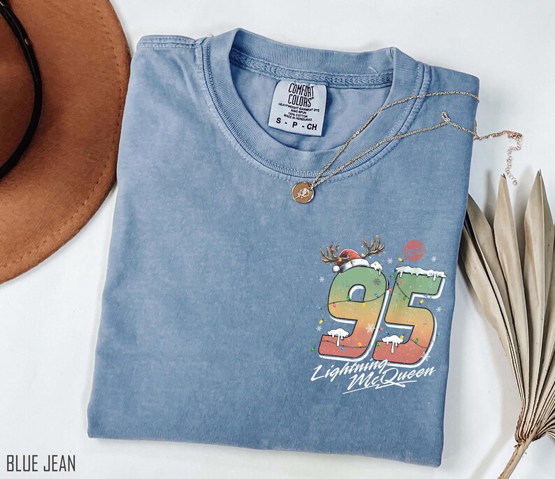 Puede incluir: Una camiseta azul vaquero con un gr&aacute;fico de Rayo McQueen con un gorro de Pap&aacute; Noel y el n&uacute;mero 95 con luces de Navidad. El texto "Lightning McQueen" est&aacute; debajo del n&uacute;mero.