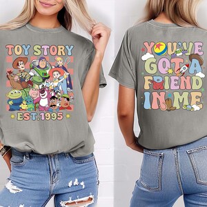 Może przedstawiać: Dwie szare koszulki z grafiką z Toy Story. Pierwsza koszulka zawiera napis "Toy Story" i obraz Woody'ego, Buzza Astrala i innych postaci z filmu. Druga koszulka zawiera napis "You've Got a Friend in Me" w kolorowej, retro czcionce.