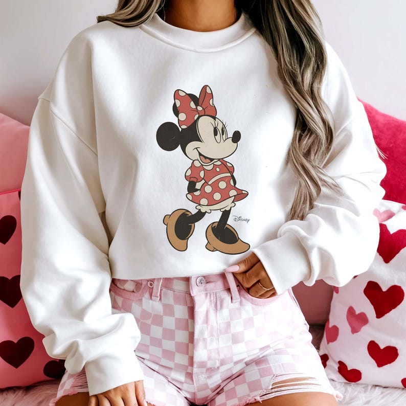 May include: A white sweatshirt featuring a Minnie Mouse graphic. Minnie is depicted in her classic outfit: a red polka-dot dress and bow, and brown shoes. The Disney logo is visible. The model is wearing pink checkered shorts.