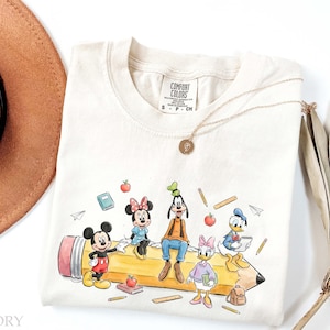 May include: An ivory t-shirt featuring a graphic print of Mickey Mouse, Minnie Mouse, Goofy, Daisy Duck, and Donald Duck sitting on a large yellow pencil. The design includes small illustrations of apples, books, rulers, and pencils. The shirt has a Comfort Colors label at the neck.