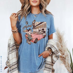May include: A light blue t-shirt featuring a vintage-style graphic of Mickey Mouse and Minnie Mouse in a pink convertible. The graphic includes the word "Mickey" and a diner scene. The shirt has a classic, casual style.