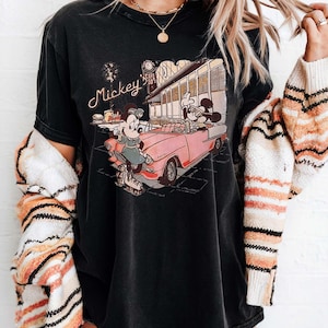 May include: Black t-shirt featuring a vintage-style graphic of Mickey and Minnie Mouse in a pink convertible. The graphic includes the text "Mickey's Cafe 50's". The shirt has a slightly faded appearance.