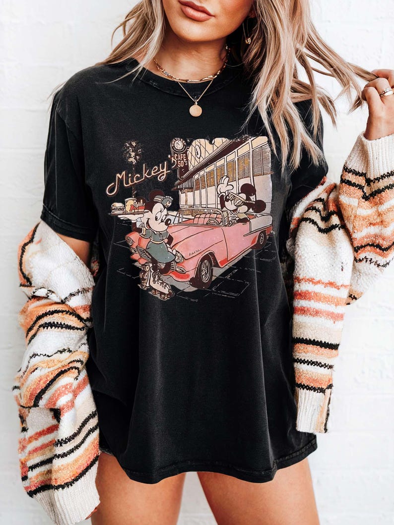 May include: Black t-shirt featuring a vintage-style graphic of Mickey and Minnie Mouse in a pink convertible. The graphic includes the text "Mickey's Cafe 50's". The shirt has a slightly faded appearance.