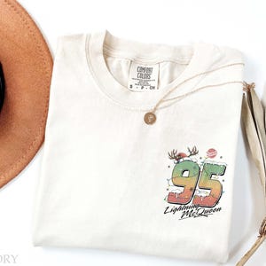 Puede incluir: Una camiseta de color crema con un gr&aacute;fico de un n&uacute;mero 95 rojo y verde con las palabras "Lightning McQueen" debajo. El gr&aacute;fico est&aacute; rodeado de nieve blanca y luces de Navidad rojas y verdes. La camiseta tiene una etiqueta que dice "Comfort Colors" y la talla es S.