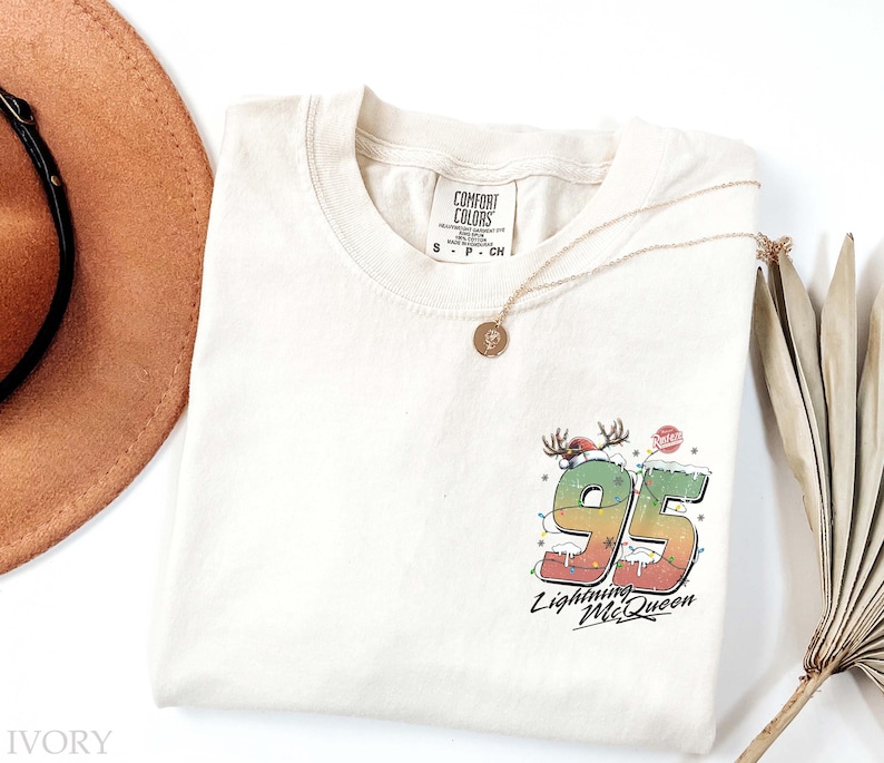 Puede incluir: Una camiseta de color crema con un gr&aacute;fico de un n&uacute;mero 95 rojo y verde con las palabras "Lightning McQueen" debajo. El gr&aacute;fico est&aacute; rodeado de nieve blanca y luces de Navidad rojas y verdes. La camiseta tiene una etiqueta que dice "Comfort Colors" y la talla es S.