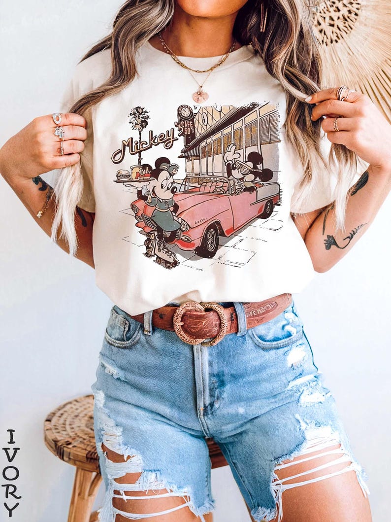 May include: Ivory-colored t-shirt featuring a vintage-style graphic of Mickey and Minnie Mouse in a pink convertible. The design includes the text "Mickey" and a retro diner scene. The shirt is paired with denim shorts and a brown belt.