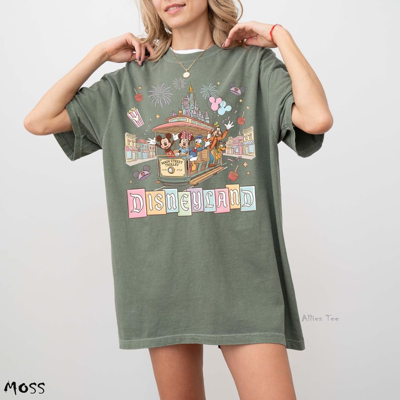 May include: Moss green t-shirt featuring a Disneyland graphic. The design includes a trolley with Mickey Mouse, Donald Duck, and Goofy, along with the Disneyland logo in pastel colors. Fireworks and other park elements are also included.