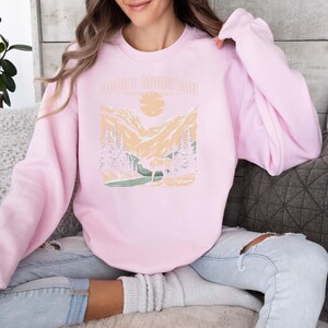 Pu&ograve; includere: Una felpa rosa con una grafica di una catena montuosa e il testo "Rocky Mountain" stampato sul davanti.