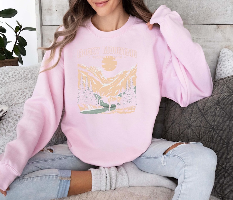 Pu&ograve; includere: Una felpa rosa con una grafica di una catena montuosa e il testo "Rocky Mountain" stampato sul davanti.