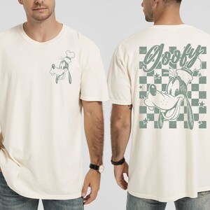 May include: Cream-colored t-shirt featuring a cartoon image of Goofy wearing a chef's hat. The front has a small image of Goofy, while the back displays a larger design with the word "Goofy" and a checkered pattern. The design is in a muted green.