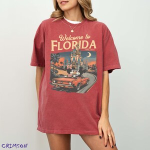 May include: Red t-shirt with a graphic featuring Mickey and Minnie Mouse in a convertible, driving towards a castle. The text "Welcome to Florida" and "Est. 1971" are displayed above the image. The shirt is a solid red color.