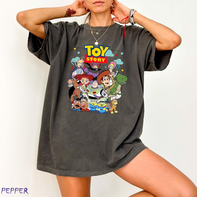 May include: Dark grey t-shirt with a vibrant graphic of characters from the animated film "Toy Story." The design includes Woody, Buzz Lightyear, Jessie, and other characters, with the words "Toy Story" in a yellow and blue font.