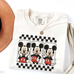May include: Ivory-colored Comfort Colors t-shirt featuring three Mickey Mouse illustrations in black and red. Each Mickey is framed within a black and white checkered border. A gold necklace and a brown hat are also visible.