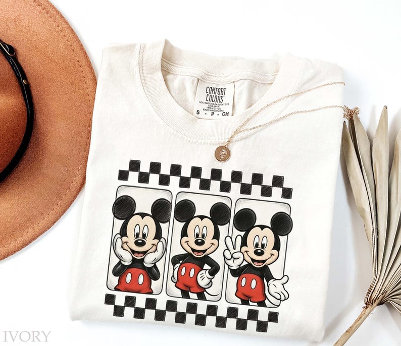 May include: Ivory-colored Comfort Colors t-shirt featuring three Mickey Mouse illustrations in black and red. Each Mickey is framed within a black and white checkered border. A gold necklace and a brown hat are also visible.