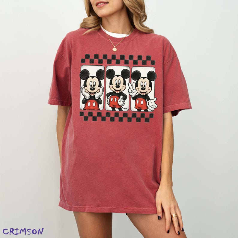 May include: A red-toned t-shirt featuring three Mickey Mouse images within black and white checkered frames. Mickey is depicted in various poses, wearing his signature red shorts and white gloves. The word "CRIMSON" is printed in the bottom left corner.