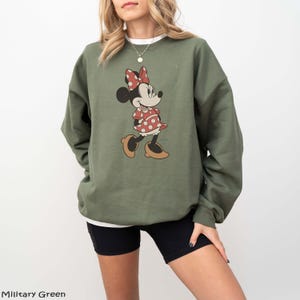 May include: An olive green sweatshirt featuring a Minnie Mouse graphic. The cartoon character is depicted in a red and white polka dot dress and bow. The sweatshirt is a relaxed fit, perfect for casual wear.