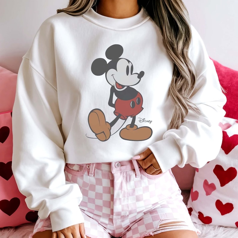 May include: A white sweatshirt featuring a graphic of Mickey Mouse in his classic pose. Mickey is depicted in black, red, and tan colors. The Disney logo is printed on the lower right side of the graphic. The sweatshirt has long sleeves.
