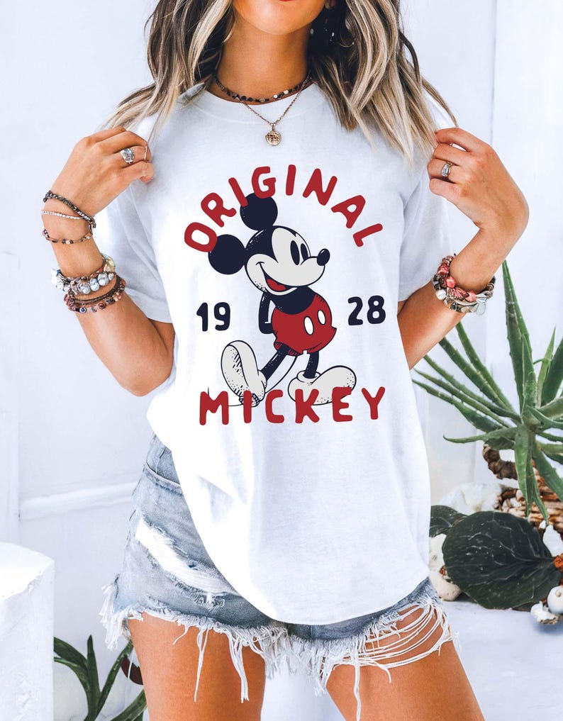 May include: White t-shirt featuring a graphic of Mickey Mouse with the text "ORIGINAL MICKEY 1928" in red and black. The design includes a smiling Mickey Mouse in his classic outfit. The shirt is a casual, everyday wear item.