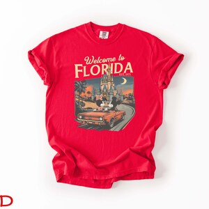 May include: A red t-shirt with a graphic that says "Welcome to Florida" and "Est. 1971". The graphic features a vintage car with Mickey and Minnie Mouse in front of a castle and palm trees.