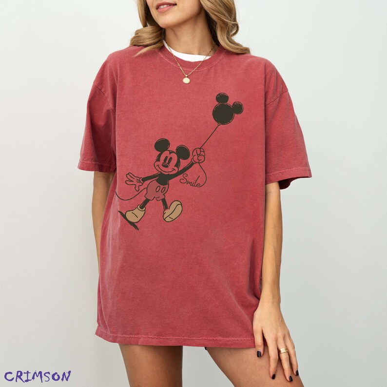 May include: Crimson red t-shirt with a black and beige Mickey Mouse graphic holding a balloon. The balloon has the word "Smile" on it. The shirt has short sleeves and a loose fit.