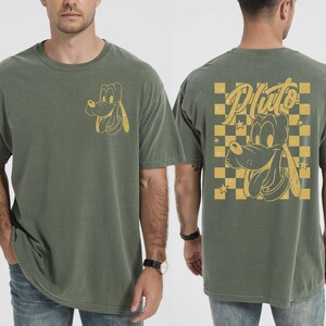 May include: Moss green t-shirt featuring a Pluto graphic. The front has a small Pluto design, while the back displays a larger Pluto image with the word "Pluto" in a stylized font, set against a yellow and black checkered background.