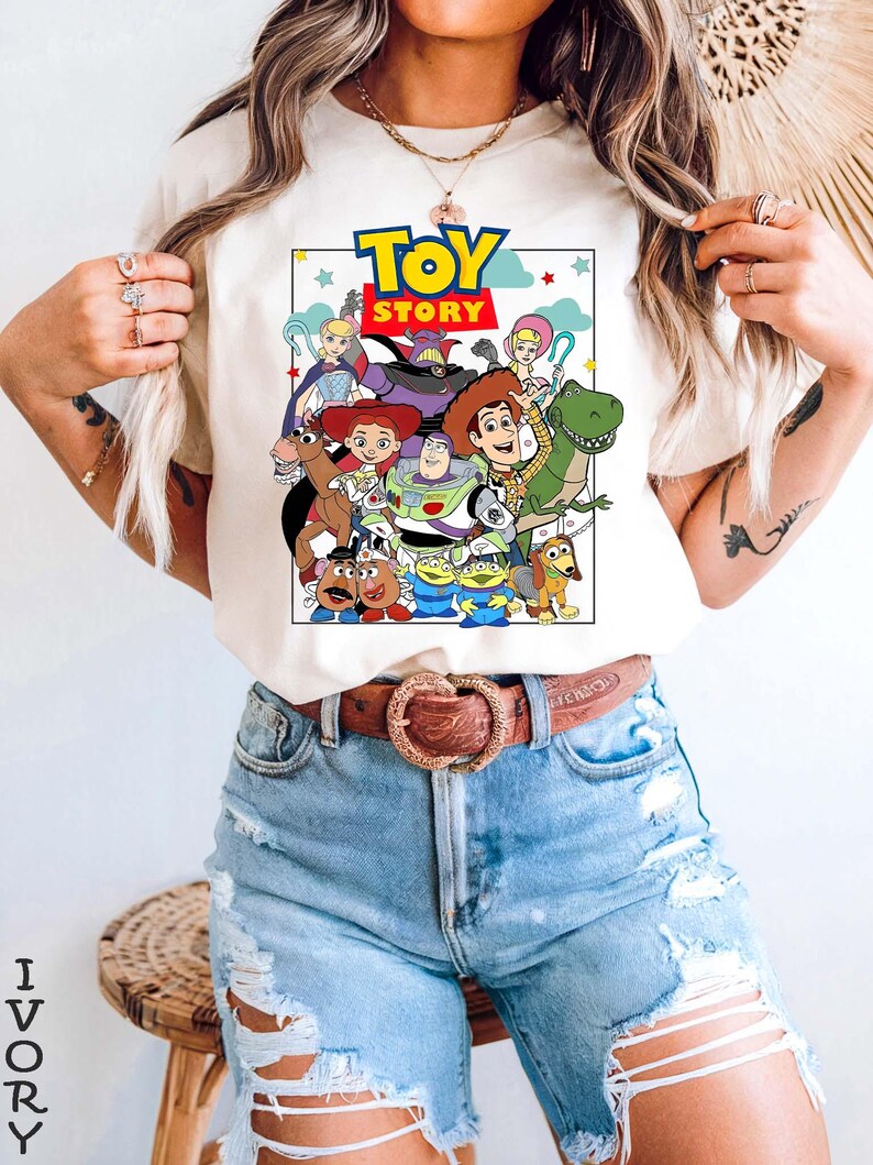 May include: Ivory t-shirt with a vibrant graphic of characters from the animated film "Toy Story." The design includes Woody, Buzz Lightyear, and other characters. The text "TOY STORY" is displayed at the top. The shirt is paired with light blue denim shorts.
