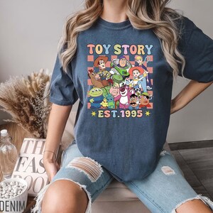 Może przedstawiać: Koszulka w kolorze granatowym z grafiką postaci z serii Toy Story. Grafika zawiera Woody'ego, Buzz Lightyeara i inne postacie. Na koszulce nadrukowany jest tekst "Toy Story Est. 1995".