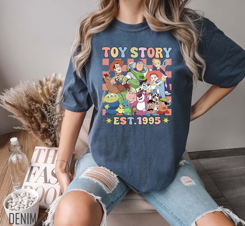 Może przedstawiać: Koszulka w kolorze granatowym z grafiką postaci z serii Toy Story. Grafika zawiera Woody'ego, Buzz Lightyeara i inne postacie. Na koszulce nadrukowany jest tekst "Toy Story Est. 1995".