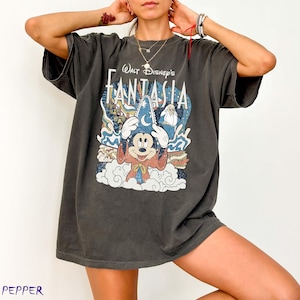 May include: Dark grey t-shirt with a Walt Disney's Fantasia graphic. The design features Mickey Mouse in a sorcerer's hat, clouds, and the film's title. The shirt has short sleeves and a loose fit.