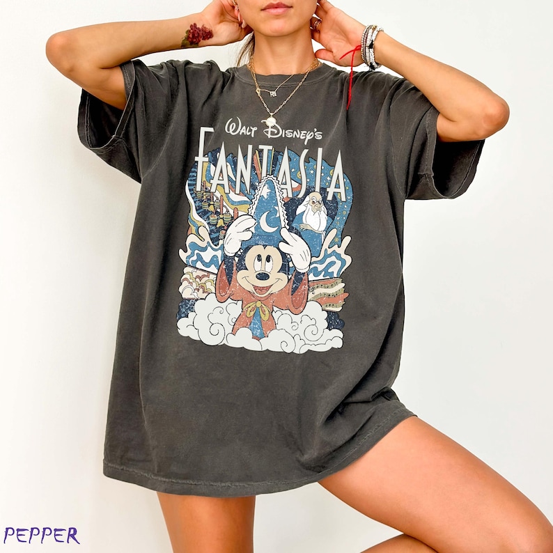 May include: Dark grey t-shirt with a Walt Disney's Fantasia graphic. The design features Mickey Mouse in a sorcerer's hat, clouds, and the film's title. The shirt has short sleeves and a loose fit.