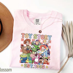 Może przedstawiać: R&oacute;żowa koszulka z grafiką postaci z Toy Story, w tym Woody'ego, Buzz Lightyeara i Jessie. Grafika jest otoczona szachownicą i tekstem "Toy Story Est. 1995".