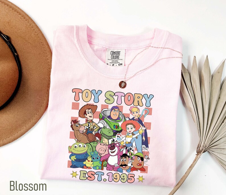 Może przedstawiać: R&oacute;żowa koszulka z grafiką postaci z Toy Story, w tym Woody'ego, Buzz Lightyeara i Jessie. Grafika jest otoczona szachownicą i tekstem "Toy Story Est. 1995".