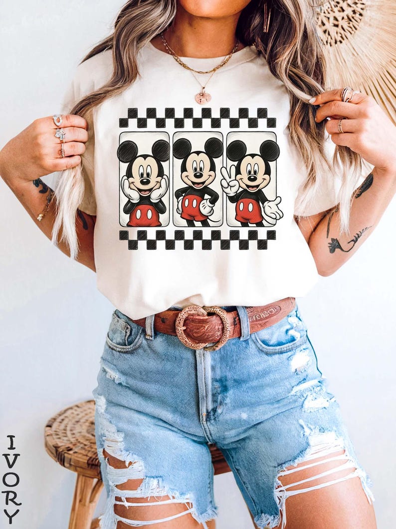 May include: Ivory-colored t-shirt featuring three Mickey Mouse illustrations within a black and white checkered border. Mickey is depicted in various poses, wearing his signature red shorts and white gloves. The shirt is paired with light blue denim shorts and a brown belt.