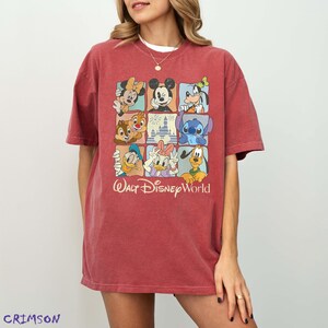 Op de afbeelding: Rood T-shirt met een rasterontwerp met Disney-personages zoals Mickey Mouse, Minnie Mouse, Goofy, Donald Duck en Stitch. De woorden "Walt Disney World" zijn in cursieve letters onder het raster gedrukt.