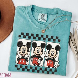 May include: Seafoam green t-shirt featuring three Mickey Mouse images in a black and white checkered frame. Mickey is wearing red shorts and a black shirt. A gold necklace with a pendant is on the shirt.