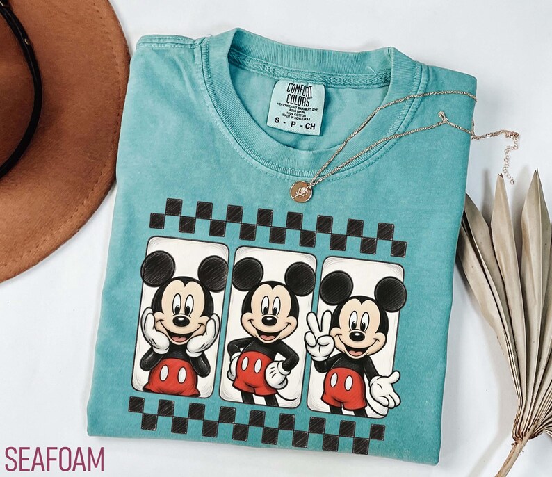May include: Seafoam green t-shirt featuring three Mickey Mouse images in a black and white checkered frame. Mickey is wearing red shorts and a black shirt. A gold necklace with a pendant is on the shirt.