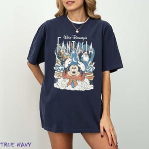 May include: A navy blue t-shirt with the words "Walt Disney's Fantasia" and a graphic of Mickey Mouse in a sorcerer's hat. The design includes clouds, stars, and characters from the film. The t-shirt has a loose fit.