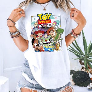 May include: White t-shirt with a vibrant graphic of characters from the animated film "Toy Story." The design includes the film's title in a blue and yellow font, surrounded by characters such as Woody, Buzz Lightyear, and Rex.