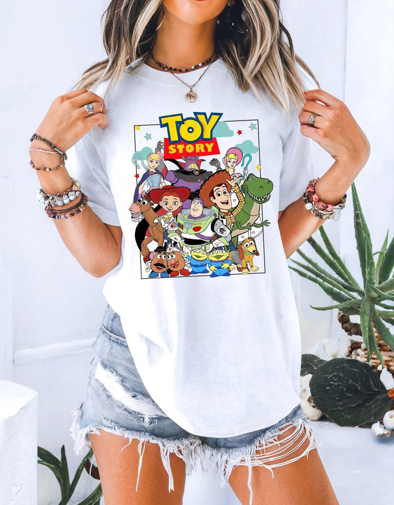 May include: White t-shirt with a vibrant graphic of characters from the animated film "Toy Story." The design includes the film's title in a blue and yellow font, surrounded by characters such as Woody, Buzz Lightyear, and Rex.