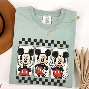 May include: A light green t-shirt featuring a graphic of Mickey Mouse in three different poses. The shirt has a black and white checkered border design. A gold necklace is draped across the shirt.