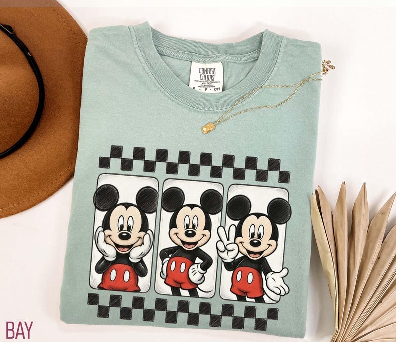 May include: A light green t-shirt featuring a graphic of Mickey Mouse in three different poses. The shirt has a black and white checkered border design. A gold necklace is draped across the shirt.