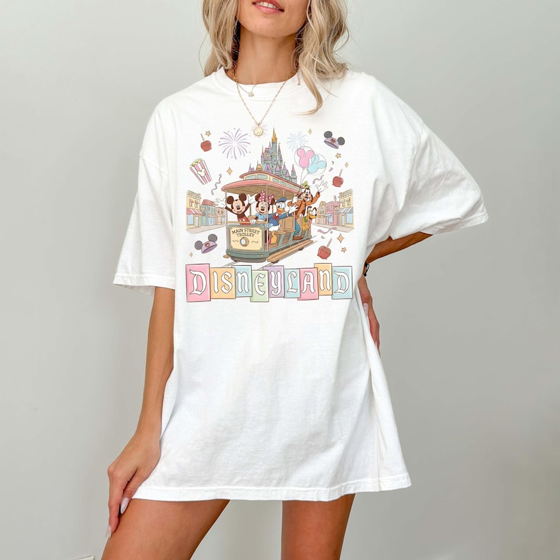 May include: White t-shirt featuring a Disneyland design. The graphic includes a trolley with Disney characters, a castle, and the word "Disneyland" in pastel colors. The shirt has short sleeves and a relaxed fit.