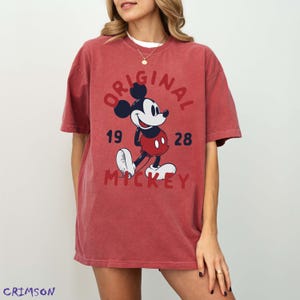 May include: Red t-shirt featuring a graphic of Mickey Mouse with the text "ORIGINAL MICKEY 1928". The design includes Mickey in his classic outfit. The shirt is a relaxed fit.