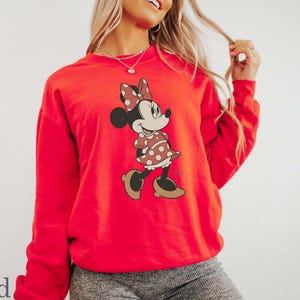 May include: Red sweatshirt featuring a Minnie Mouse graphic. The cartoon character is depicted in a polka-dot dress and bow. The word "red" is printed in the bottom left corner. The sweatshirt has long sleeves and a crew neck.