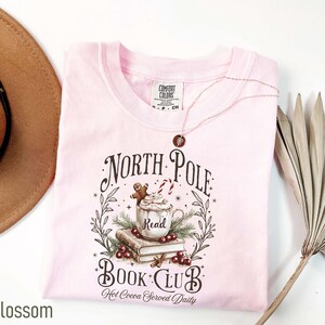 K&ouml;nnte beinhalten: Hellrosa T-Shirt mit dem Schriftzug "North Pole Book Club" und einer Grafik einer Tasse hei&szlig;e Schokolade, B&uuml;cher und einem Lebkuchenmann. Das Shirt ist aus Comfort Colors Material.