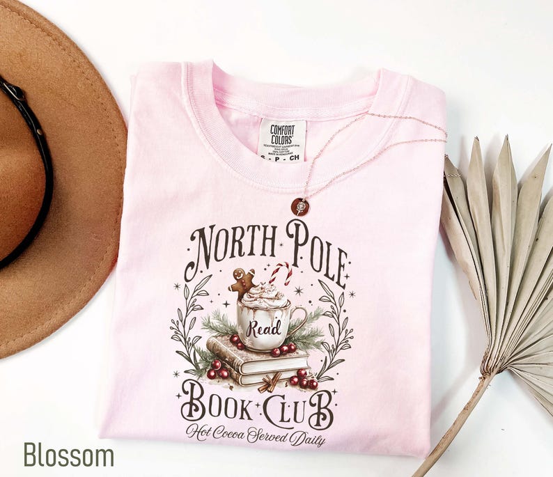 K&ouml;nnte beinhalten: Hellrosa T-Shirt mit dem Schriftzug "North Pole Book Club" und einer Grafik einer Tasse hei&szlig;e Schokolade, B&uuml;cher und einem Lebkuchenmann. Das Shirt ist aus Comfort Colors Material.