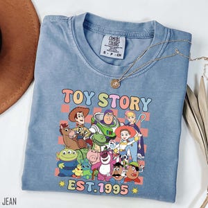 Może przedstawiać: Jasnoniebieska koszulka z przetartym efektem, przedstawiająca kolorową grafikę postaci z filmu Toy Story. Grafika zawiera tekst "Toy Story Est. 1995".