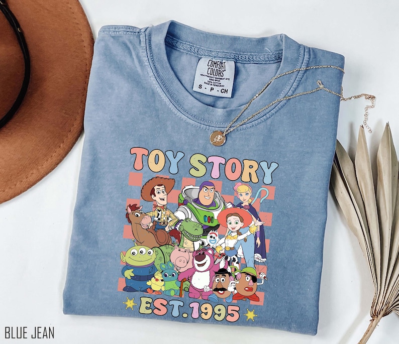 Może przedstawiać: Jasnoniebieska koszulka z przetartym efektem, przedstawiająca kolorową grafikę postaci z filmu Toy Story. Grafika zawiera tekst "Toy Story Est. 1995".