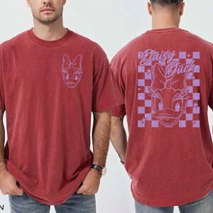 May include: Crimson red t-shirt featuring a Daisy Duck graphic. The front of the shirt has a small Daisy Duck outline, while the back displays a larger design with the words "Daisy Duck" and a checkered pattern. The shirt is a relaxed fit.
