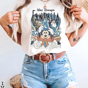 May include: Ivory t-shirt with a vintage graphic of Mickey Mouse from Disney's Fantasia. The design includes the text "Walt Disney's Fantasia" in a stylized font, with Mickey wearing a sorcerer's hat. The shirt is worn with denim shorts.